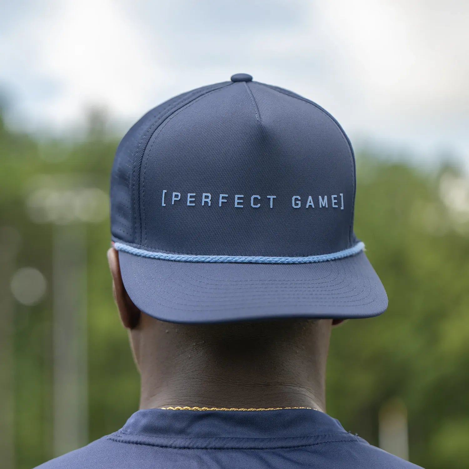 Person wearing a blue cap with 'PERFECT GAME' text, blurred green background